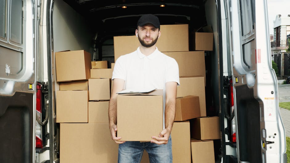 Two movers from Man with Van Crayford are engaged in a home relocation process, loading large cardboard boxes into a spacious van with an open rear door on a clear day. The man, wearing a white shirt,
