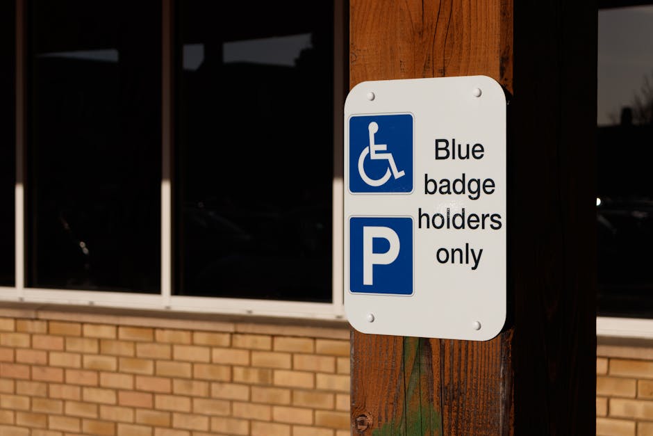 A white rectangular parking sign attached to a wooden post outside a building, indicating parking for blue badge holders only with a blue wheelchair symbol and a blue parking 'P' symbol. The sign is positioned near a brick wall and large window panes. In the background, reflections of parked cars are visible through the darkened glass. The scene suggests a designated accessible parking area, relevant to home relocation services by Man with Van Crayford, where properly designated parking spaces are important for efficient furniture transport and moving logistics during house removals or property moves within the Crayford area.