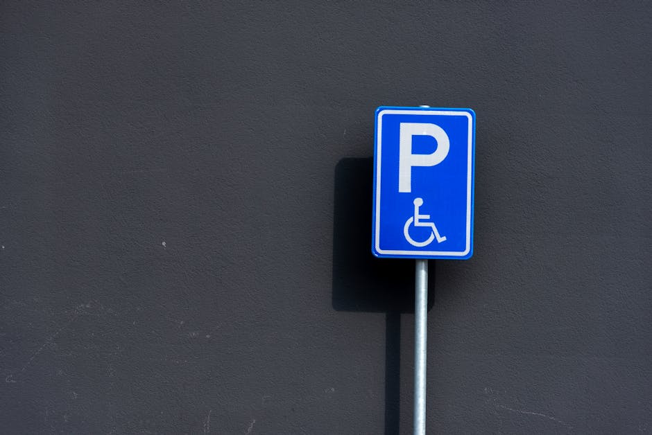 A blue rectangular parking permit sign with a white 'P' and a wheelchair symbol is mounted on a metal pole against a dark grey textured wall. The sign indicates designated parking for individuals with disabilities. The shadow of the sign is cast onto the wall, suggesting daylight. This signage is typically found outside residential or commercial properties, useful during home relocation or furniture transport to ensure accessible parking arrangements. The sign's location in a well-lit outdoor area indicates proximity to a property where a house removal or moving service, such as those provided by Man with Van Crayford, may be operating, supporting efficient loading and unloading processes involved in packing, moving, and home relocation activities.
