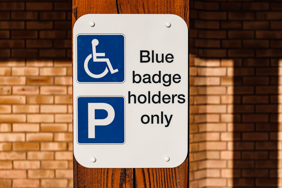A white rectangular parking sign attached to a wooden post outside a building, indicating parking for blue badge holders only with a blue wheelchair symbol and a blue parking 'P' symbol. The sign is positioned near a brick wall and large window panes. In the background, reflections of parked cars are visible through the darkened glass. The scene suggests a designated accessible parking area, relevant to home relocation services by Man with Van Crayford, where properly designated parking spaces are important for efficient furniture transport and moving logistics during house removals or property moves within the Crayford area.