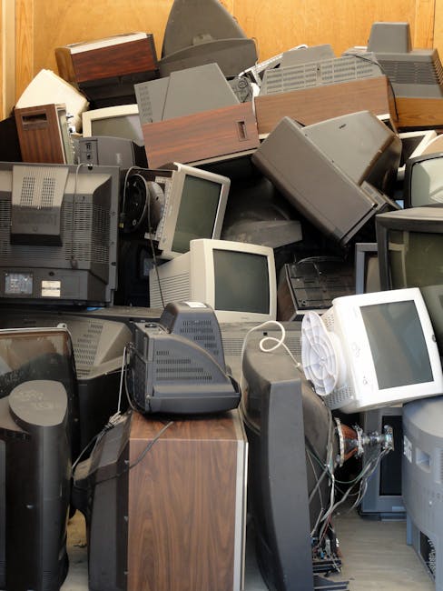 A large pile of old electronic devices and equipment, including vintage CRT computer monitors, box-shaped speakers, and various types of computer peripherals, stacked haphazardly on a wooden floor inside an indoor space. Some items are stacked on top of each other, with visible cords and cables tangled around the equipment. In the background, there is a wooden wall or panel, indicating an interior setting likely used for storage or disposal. The space appears cluttered, reflecting the process of preparing items for removal or recycling as part of a home relocation or clearance service. The image is associated with Man with Van Crayford's removals and waste disposal services, illustrating the type of furniture and electronic waste handled during a bulky waste move.