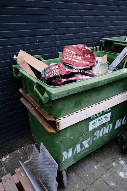 A green residential waste bin filled with red and white bags containing garden waste and outdoor equipment, positioned on a paved surface next to a dark brick wall. The lid of the bin is open, revealing the contents inside, including additional cardboard boxes and plastic packaging materials. Partly visible underneath the green bin is a white waste container with green labels, and nearby are a metal ramp and some wooden panels on the ground, indicating a recent or upcoming house removal or relocation process facilitated by a professional removal service such as Man with Van Crayford. The scene suggests a typical home clearance or packing task involving outdoor waste disposal as part of furniture transport or moving logistics.