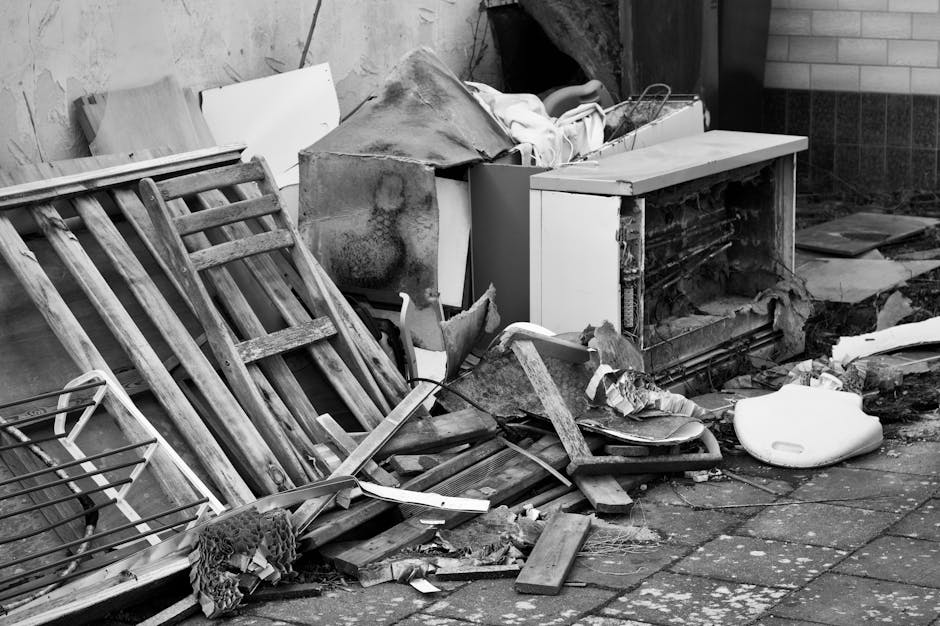 A large pile of old electronic devices and equipment, including vintage CRT computer monitors, box-shaped speakers, and various types of computer peripherals, stacked haphazardly on a wooden floor inside an indoor space. Some items are stacked on top of each other, with visible cords and cables tangled around the equipment. In the background, there is a wooden wall or panel, indicating an interior setting likely used for storage or disposal. The space appears cluttered, reflecting the process of preparing items for removal or recycling as part of a home relocation or clearance service. The image is associated with Man with Van Crayford's removals and waste disposal services, illustrating the type of furniture and electronic waste handled during a bulky waste move.
