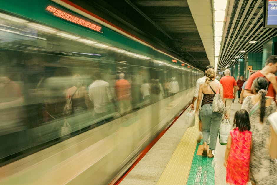 People standing on a platform at Crayford Station in the UK, waiting to board a fast-moving train visible with blurred motion effects reflecting passengers inside. The scene shows a mix of adults and children, some carrying shopping bags and backpacks, positioned near the platform's edge with tactile paving and safety markings. The station's green and metal ceiling panels, along with the illuminated signage and doors of the train, are visible in the background. This setting illustrates the typical environment for a home relocation preparation or a commuter transfer, emphasizing efficient, timely transport processes associated with professional removals services such as those offered by Man with Van Crayford, particularly relevant for moving logistics and furniture transport related to house moves near Crayford station.