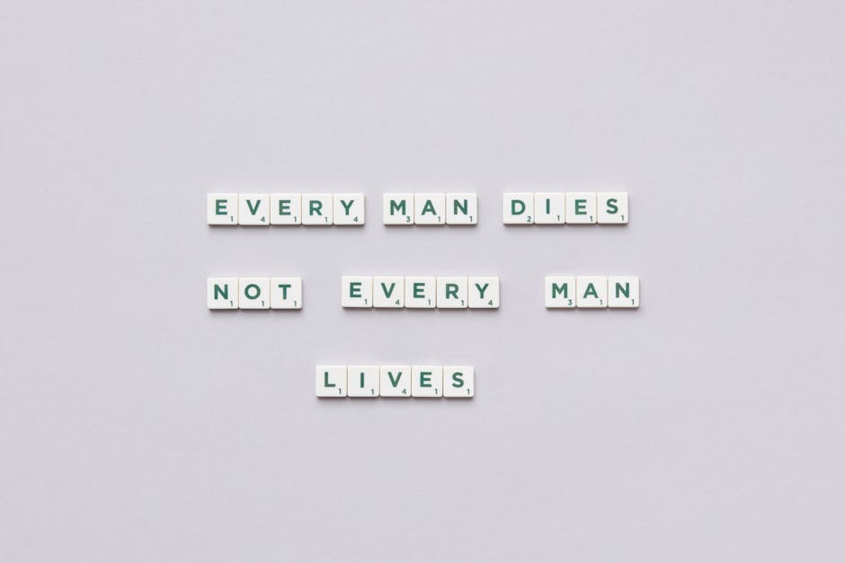 A flat lay image of white letter tiles with black and green letters, arranged to spell out the message 'EVERY MAN DIES NOT EVERY MAN LIVES' on a plain light grey background. The tiles are organized in a staggered layout, with the first line reading 'EVERY MAN DIES', the second line 'NOT EVERY MAN', and the third line 'LIVES'. The setting is minimalistic, emphasizing the message with clear, evenly spaced tiles. This visual can relate to themes of life and mortality, paralleling the strategic planning involved in home relocation and furniture transport, which are key services offered by Man with Van Crayford, particularly in the context of efficient packing and loading during house removals.