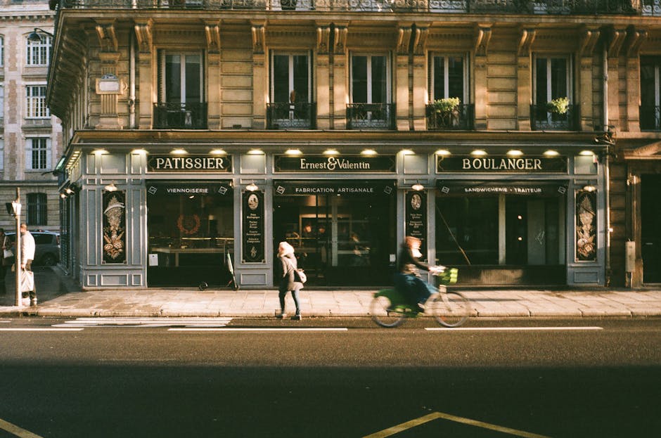 The image depicts the façade of a traditional Parisian-style building with a ground-floor bakery or pastry shop named 'Ernest & Valentin,' which is part of a house removal or furniture transport operation. The storefront features large windows with decorative painted panels illustrating baked goods, flanked by black signage with gold lettering indicating 'Pâtissier,' 'Boulanger,' and 'fabrication artisanale.' Above the shop, the building has multiple floors with tall windows, some with window boxes containing green plants, suggesting residential or office spaces. In front of the shop, the pavement is visible, with pedestrians walking along the sidewalk—one person is walking, and another appears to be using a bicycle equipped with a basket, in motion, creating a slight blur. The street traffic includes tram tracks or road markings, indicating an urban setting suitable for home relocation and moving services. The scene is illuminated by street lighting or ambient evening light, emphasizing the shop's exterior details and the urban environment where furniture or household items might be being moved via professional removals provided by companies like Man with Van Crayford, supporting packing and moving logistics.