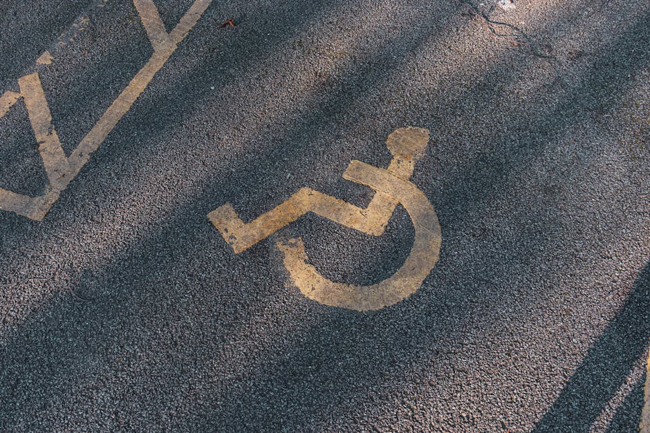 Close-up of a disabled parking bay symbol painted yellow on a dark asphalt surface outside a property. The symbol depicts a stylized figure sitting in a wheelchair, with the head, arms, and legs clearly outlined. The surrounding asphalt shows signs of wear with some texture and minor cracks. The image suggests an accessible parking area adjacent to a bungalow or house entrance, with the parking space potentially used during a home relocation or furniture transport process. This visual is relevant to house removals and moving services provided by Man with Van Crayford, especially when considering parking access challenges during a local move or home relocation in Crayford, UK.
