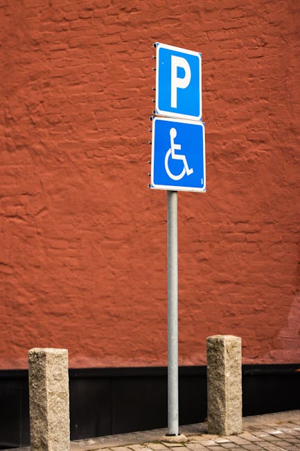 Image showing two blue parking signs mounted on a metal pole against a red brick wall. The upper sign indicates general parking with a white 'P' symbol, while the lower sign displays a wheelchair symbol designating accessible parking. The signs are positioned on a paved sidewalk with two stone bollards at the base of the pole, creating a boundary for vehicle or pedestrian access. This scene highlights urban parking regulations, which may be relevant for people arranging moves with Man with Van Crayford, especially when considering parking permits or accessible loading zones during home removals or furniture transport in Crayford.