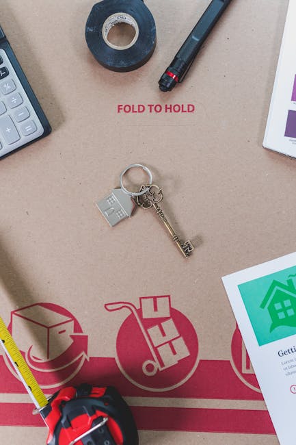 Close-up image of a cardboard moving box with red printed instructions for folding and handling, situated on a flat surface during a house relocation process. The box features partially visible icons indicating packing and transport steps. On top of the box, there is a set of metallic keys with a house-shaped keychain, a calculator, a roll of black adhesive tape, a black marker pen, and a diary or checklist with colour-coded icons and text related to moving procedures. To the side, a yellow measuring tape and a portion of a white tablet displaying a green house icon and information about a move are also visible. The environment suggests an organized packing or planning stage of furniture transport, with items indicative of a professional removals service such as Man with Van Crayford. The lighting is natural, highlighting the details of the packing materials and tools used in the relocation process.
