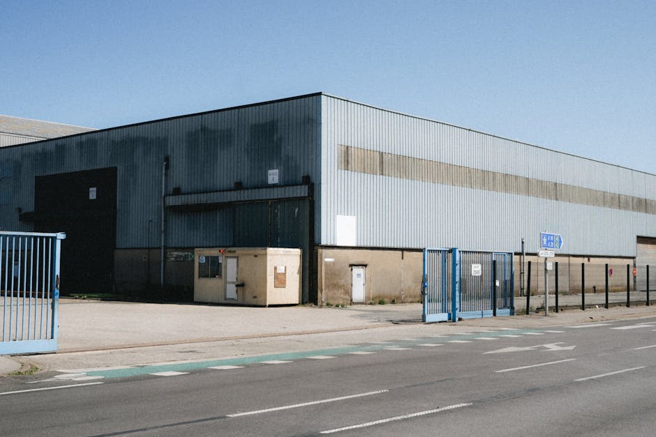 Exterior view of a large industrial warehouse on Crayford Industrial Estate under clear blue sky, with corrugated metal cladding in shades of grey and beige. The building features large black roller shutter doors and a smaller personnel door, all positioned along the front facade. In front of the warehouse, there is a paved area with several medium-sized cardboard boxes and a wooden-framed mirror packed with protective plastic wrap, ready for transportation. A blue metal pedestrian gate and a similar gate for vehicle access are open, leading onto a pavement and a nearby road marked with white dashed and solid lines, indicating a parking or loading zone. In the foreground, a man from Man with Van Crayford, specializing in removals, is seen lifting a covered piece of furniture, possibly a wooden table or desk, onto a dolly or trolley to facilitate loading into a van parked just outside the premises. The scene captures the process of packing and furniture transport during an office relocation case study at Crayford Industrial Estate, highlighting professional moving services in action.