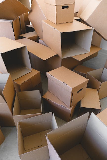 Close-up image of a cardboard moving box with red printed instructions for folding and handling, situated on a flat surface during a house relocation process. The box features partially visible icons indicating packing and transport steps. On top of the box, there is a set of metallic keys with a house-shaped keychain, a calculator, a roll of black adhesive tape, a black marker pen, and a diary or checklist with colour-coded icons and text related to moving procedures. To the side, a yellow measuring tape and a portion of a white tablet displaying a green house icon and information about a move are also visible. The environment suggests an organized packing or planning stage of furniture transport, with items indicative of a professional removals service such as Man with Van Crayford. The lighting is natural, highlighting the details of the packing materials and tools used in the relocation process.