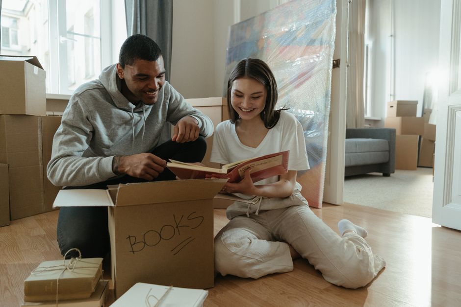 A man and a woman, both smiling, sit on the wooden floor of an interior space surrounded by packed cardboard boxes, some labelled with the word 'BOOKS.' The man, wearing a grey hoodie and dark trousers, holds a book, while the woman, dressed in a white shirt and light-colored pants, looks at the book and points at something inside it. They appear to be engaged in organizing or reviewing their belongings as part of a home relocation process. Behind them, a large wrapped item or piece of furniture is visible, along with a sofa and windows allowing natural light to illuminate the room. The scene depicts the loading process typical of professional house removals, with packaging materials and furniture prepared for transport by a removals company such as Man with Van Crayford, highlighting packing, moving, and furniture transport activities during an emergency or same-day move.