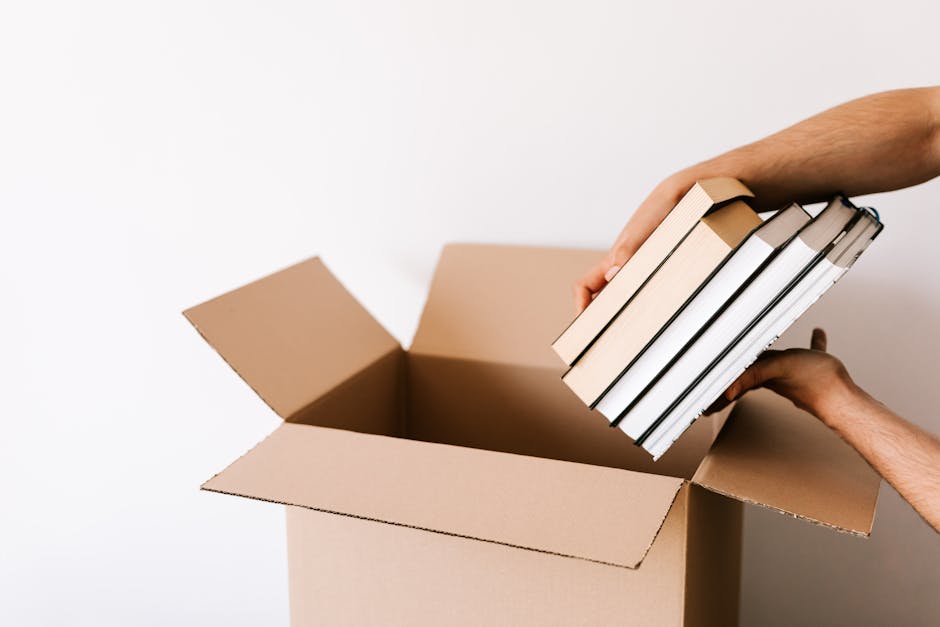 A man and a woman, both smiling, sit on the wooden floor of an interior space surrounded by packed cardboard boxes, some labelled with the word 'BOOKS.' The man, wearing a grey hoodie and dark trousers, holds a book, while the woman, dressed in a white shirt and light-colored pants, looks at the book and points at something inside it. They appear to be engaged in organizing or reviewing their belongings as part of a home relocation process. Behind them, a large wrapped item or piece of furniture is visible, along with a sofa and windows allowing natural light to illuminate the room. The scene depicts the loading process typical of professional house removals, with packaging materials and furniture prepared for transport by a removals company such as Man with Van Crayford, highlighting packing, moving, and furniture transport activities during an emergency or same-day move.