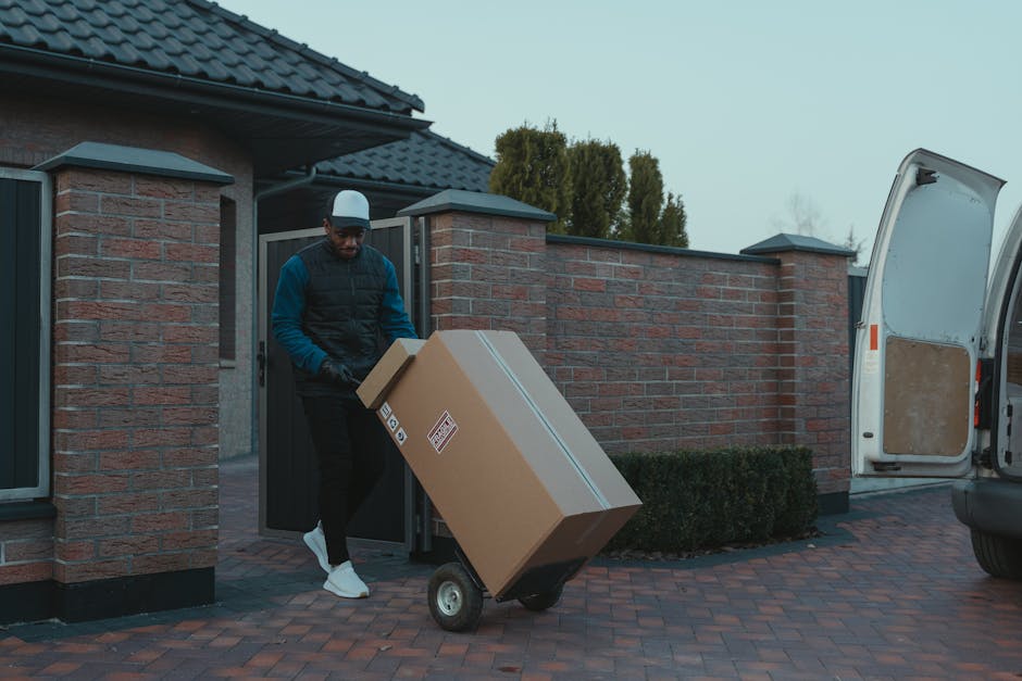 A man wearing a baseball cap, black vest, blue long-sleeve shirt, and white sneakers is pushing a black hand truck loaded with a large cardboard box secured with plastic straps during a home relocation. He is standing outside a residential property with a brick wall, a paved driveway, and a partially open white cargo van parked nearby, which has a beige interior panel visible through the open rear door. The scene takes place in daytime, with trees and a cloudy sky in the background. The man with Van Crayford is involved in furniture transport and packing as part of a professional removals service, carefully handling the loading process on the driveway, which is clean and well-maintained.