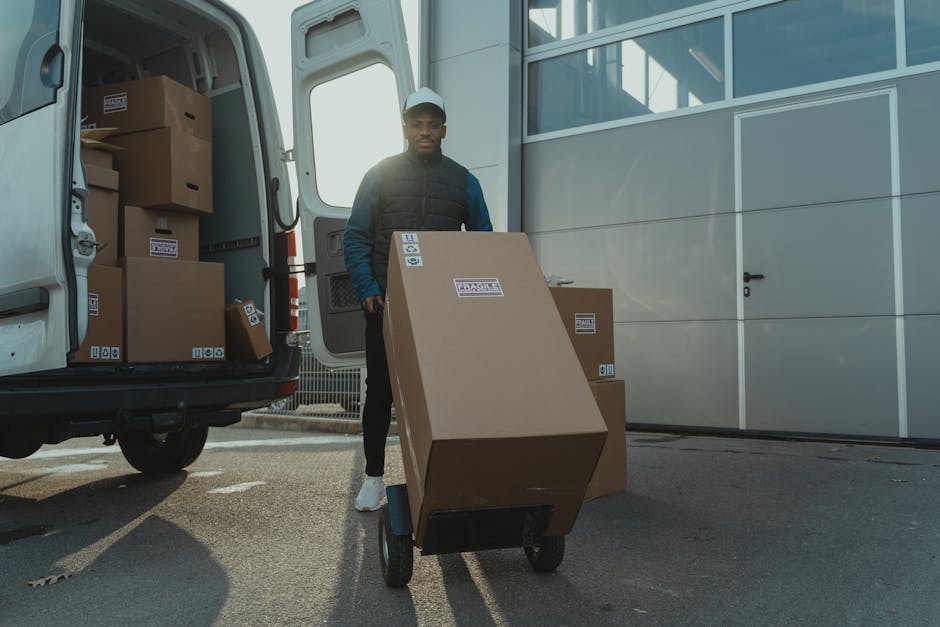 A man wearing a dark vest, blue sleeves, and a white cap is standing outside a white moving van, holding the handle of a hand truck loaded with a large cardboard box labeled with packing and handling symbols. The van's side door is open, revealing several other cardboard boxes stacked inside on the vehicle's floor. The boxes are sealed with packing tape, some are stacked on top of each other, and a few are leaning against the interior walls of the van. The loading area is on a paved surface, adjacent to the van, with the background showing a modern building with a grey metal facade and large glass windows. The image captures the process of home relocation, with the efficient movement of packed boxes towards the vehicle for transportation, typical of professional removals services such as those offered by Man with Van Crayford.