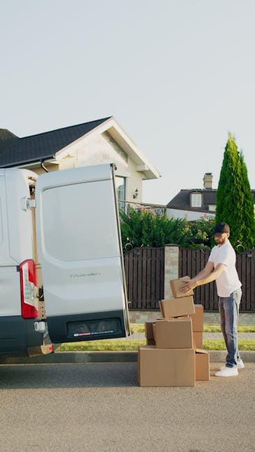 A man wearing a baseball cap, black vest, blue long-sleeve shirt, and white sneakers is pushing a black hand truck loaded with a large cardboard box secured with plastic straps during a home relocation. He is standing outside a residential property with a brick wall, a paved driveway, and a partially open white cargo van parked nearby, which has a beige interior panel visible through the open rear door. The scene takes place in daytime, with trees and a cloudy sky in the background. The man with Van Crayford is involved in furniture transport and packing as part of a professional removals service, carefully handling the loading process on the driveway, which is clean and well-maintained.