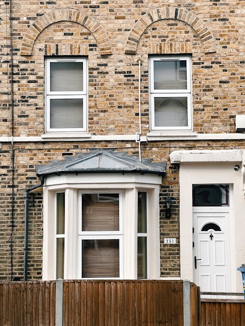A brick-built terraced house with two levels, featuring two white-framed sash windows on the upper floor, each with closed blinds. Below, on the ground floor, there is a bay window with three sections, also with closed blinds, topped by a small, sloped grey roof. To the right of the bay window, a white exterior door with a semi-circular window at the top and a black handle is visible. A black downpipe runs vertically beside the left sash window, and white pipes run horizontally along the façade at ground level. In front of the house, a wooden fence partially obscures the lower part of the bay window. The setting appears to be during the daytime, with natural lighting illuminating the stone and brickwork. This image exemplifies a typical residential property prepared for a home relocation or furniture transport, aligning with the services provided by Man with Van Crayford during house removals and packing processes.