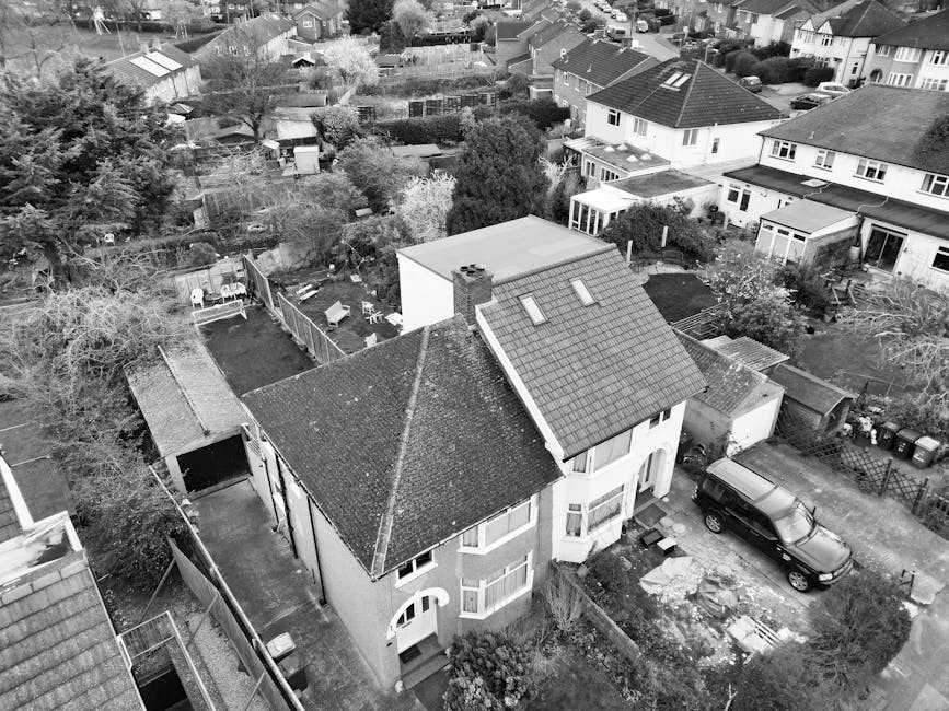 An aerial black-and-white photograph showing a house removal process at a two-storey semi-detached property in a residential area. The image captures the front of the house with a driveway where a large dark vehicle, likely a removal van, is parked. Several cardboard boxes, wrapped furniture items, and packing materials are visible on the driveway and surrounding garden areas. The property features a tiled roof with three skylights, a front porch, and large windows. The driveway is partially cleared, and some packing items are arranged nearby, indicating an ongoing packing and moving process. Adjacent houses with gardens, trees, and fenced yards fill the background, illustrating the community setting. The scene is lit with natural daylight, and the image reflects the logistics involved in house removals and furniture transport carried out by Man with Van Crayford as part of their professional relocation services.